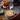 Close-up of hands pouring milk from a stainless steel pitcher into a cup of pumpkin spice latte, creating latte art on top. The background features a small orange pumpkin, cinnamon sticks, and wooden bowls of pumpkin seeds and spices, all on a grey textured surface. The image evokes a sense of warmth and autumnal coziness.
