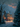 A picturesque painting depicts a serene winter night with soft snowfall. A family of four, consisting of two adults and two children, walks along a winding, snow-covered path towards a beautifully illuminated, large Victorian-style house adorned with festive lights and Christmas trees. Snow-covered evergreens and bare trees frame the scene, and towering mountains are visible in the distance under a twilight sky with warm orange and pink hues.
