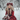 A young blonde woman with piercing blue eyes and wavy hair kneels in a snow-covered forest, gazing intently forward. She is dressed in a vibrant red coat with a fur collar and an elaborate red floral and berry crown, holding a bouquet of white flowers and red berries. Gentle snowflakes fall around her, contributing to the magical winter wonderland setting with blurred, snow-laden trees in the background.