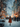 A young girl with long, wavy blonde hair and a vibrant burnt orange winter coat and beanie walks away from the viewer down a snow-covered street. The scene is illuminated by numerous soft, glowing orange and yellow string lights decorating bare tree branches and distant buildings, with countless blurred snowflakes falling, creating a magical, dreamlike winter atmosphere.