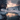 A vertical shot of a serene winter landscape at sunset. A partially frozen stream reflects warm orange and pink sky colors, winding through fresh white snow. In the midground, a small, dark-roofed house is nestled among snow-covered evergreen and bare trees, with distant snowy mountains under a dramatic, cloudy sky.