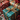 A close-up vertical shot of several Christmas presents beautifully arranged on a textured light-colored lace fabric. In the foreground, a teal gift with gold snowflake patterns and a sparkling gold ribbon bow is prominently in focus. Behind it, other gifts wrapped in red patterned paper and gold and brown striped paper are partially visible. The scene is adorned with green fir branches, red and white striped candy canes, and a warm, blurred background featuring bokeh lights and ornaments.