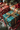 A close-up vertical shot of several Christmas presents beautifully arranged on a textured light-colored lace fabric. In the foreground, a teal gift with gold snowflake patterns and a sparkling gold ribbon bow is prominently in focus. Behind it, other gifts wrapped in red patterned paper and gold and brown striped paper are partially visible. The scene is adorned with green fir branches, red and white striped candy canes, and a warm, blurred background featuring bokeh lights and ornaments.