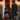 A man and two young girls in Halloween costumes stand on a porch. The girls wear witch hats and dresses, while the man wears a black coat and hat. They are facing the open doorway of a house, illuminated by warm interior lighting, holding pumpkin-shaped candy buckets and are about to go trick-or-treating.