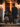 A man and two young girls in Halloween costumes stand on a porch. The girls wear witch hats and dresses, while the man wears a black coat and hat. They are facing the open doorway of a house, illuminated by warm interior lighting, holding pumpkin-shaped candy buckets and are about to go trick-or-treating.