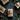 A top-down flat lay features four rustic holiday gifts nestled among fresh green fir branches, pinecones, and decorative woven ornaments, all set against a dark gray textured background. Three gifts are wrapped in natural kraft paper, and one in burlap, each tied with twine and accented with a small evergreen sprig. The composition creates a cozy and festive atmosphere with a focus on natural, earthy elements.