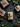 A top-down flat lay features four rustic holiday gifts nestled among fresh green fir branches, pinecones, and decorative woven ornaments, all set against a dark gray textured background. Three gifts are wrapped in natural kraft paper, and one in burlap, each tied with twine and accented with a small evergreen sprig. The composition creates a cozy and festive atmosphere with a focus on natural, earthy elements.