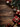 A top-down view of various Christmas decorations and gifts arranged on a dark brown wooden surface with visible grain. The festive arrangement includes red and gold baubles, natural pine cones, several wrapped presents in red and craft paper with ribbons, and green pine branches, all framing an empty central space. The image evokes a warm, festive, and traditional holiday mood, ideal for seasonal greetings.