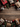A top-down flat lay shows various Christmas decorations arranged on a rustic wooden background. Gift boxes wrapped in red snowflake paper, kraft paper, and textured gold are intermingled with natural pine cones, green evergreen branches, shiny red baubles, and white snowflake ornaments, framing the top and bottom edges of the image. The central area features empty, weathered wood planks lightly dusted with artificial snow, providing clear space for text or additional design elements.