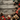 A top-down view of an assortment of beautifully wrapped Christmas gifts and festive decorations scattered around the edges of a rustic, weathered wooden table. The gifts are wrapped in red, kraft, and dark teal paper, adorned with gold ribbons, pine sprigs, and red berries, interspersed with natural pinecones, gold stars, and shiny gold and teal ornaments. The arrangement creates a central empty space, ideal for text overlay or branding.
