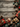 A top-down view of an assortment of beautifully wrapped Christmas gifts and festive decorations scattered around the edges of a rustic, weathered wooden table. The gifts are wrapped in red, kraft, and dark teal paper, adorned with gold ribbons, pine sprigs, and red berries, interspersed with natural pinecones, gold stars, and shiny gold and teal ornaments. The arrangement creates a central empty space, ideal for text overlay or branding.