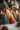 A close-up shot of several lit, decorative candles shaped like evergreen trees on a textured white tablecloth. In the foreground, a dark teal candle and an ombre orange-to-brown candle are prominently displayed with flickering flames. Behind them, additional tree-shaped candles in creamy white, sage green, and a taller brown pillar candle contribute to a warm, festive scene, complemented by blurred pinecones and a decorated Christmas tree with glowing lights in the background.