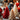 A vertical, warm-toned close-up of three lit Christmas tree-shaped candles on a textured grey surface. In the foreground, a detailed red tree candle and a cream-colored, slightly glittery tree candle burn brightly. Various Christmas decorations like pinecones, golden stars, and red baubles are scattered around, while a blurred background of warm, golden bokeh lights creates a festive and cozy atmosphere.