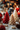 A vertical, warm-toned close-up of three lit Christmas tree-shaped candles on a textured grey surface. In the foreground, a detailed red tree candle and a cream-colored, slightly glittery tree candle burn brightly. Various Christmas decorations like pinecones, golden stars, and red baubles are scattered around, while a blurred background of warm, golden bokeh lights creates a festive and cozy atmosphere.
