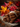 A close-up shot shows a stack of cinnamon sticks tied together with twine sitting on a square piece of burlap material. The cinnamon sticks are dark brown and have visible curls at the ends. Behind the cinnamon sticks, the background is blurred, showing out-of-focus colorful autumn leaves and red berries.