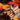 Close-up shot of a bundle of cinnamon sticks tied with a rustic twine, arranged on a piece of burlap. A small yellow autumn leaf rests nearby on a dark wooden surface. Blurred red and orange leaves are visible in the background, creating a warm, autumnal atmosphere.