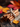Close-up shot of a bundle of cinnamon sticks tied with a rustic twine, arranged on a piece of burlap. A small yellow autumn leaf rests nearby on a dark wooden surface. Blurred red and orange leaves are visible in the background, creating a warm, autumnal atmosphere.