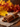 A close-up shot depicts a bundle of brown cinnamon sticks, tied with a natural twine, resting on a square of coarse burlap fabric atop a dark wooden surface. The background features blurred autumn leaves in warm yellow, orange, and red tones. Loose cinnamon powder is subtly scattered beside the burlap.