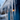 A close-up, low-angle shot of numerous translucent icicles hanging from a roof's eaves. The foreground icicles are in sharp focus, showing their clear, glistening forms, while the background is softly blurred, revealing a deep blue house wall, a light blue sky, and a warm, glowing light from inside the house.