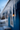 A close-up, low-angle shot of numerous translucent icicles hanging from a roof's eaves. The foreground icicles are in sharp focus, showing their clear, glistening forms, while the background is softly blurred, revealing a deep blue house wall, a light blue sky, and a warm, glowing light from inside the house.