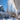A close-up, upward view of multiple long, clear icicles hanging from a white roof eave. The icicles are brightly lit and appear to be melting slightly, with some water droplets visible. In the heavily blurred background, a vibrant blue sky, the peak of a blue house, and snow-covered grounds with subtle shadows are visible under bright sunlight.