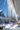 A close-up, upward view of multiple long, clear icicles hanging from a white roof eave. The icicles are brightly lit and appear to be melting slightly, with some water droplets visible. In the heavily blurred background, a vibrant blue sky, the peak of a blue house, and snow-covered grounds with subtle shadows are visible under bright sunlight.