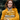 A young white woman with long, wavy red hair and freckles smiles warmly, looking directly at the viewer. She is wearing a mustard yellow textured coat, a patterned neutral-toned scarf, and gold hoop earrings, holding a stack of four elaborately wrapped gold and white gifts in her arms. In the blurred background are the deep green needles of a fir tree.