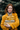 A young white woman with long, wavy red hair and freckles smiles warmly, looking directly at the viewer. She is wearing a mustard yellow textured coat, a patterned neutral-toned scarf, and gold hoop earrings, holding a stack of four elaborately wrapped gold and white gifts in her arms. In the blurred background are the deep green needles of a fir tree.
