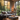 The image shows a modern living room with a large window showcasing autumn foliage. A gray sofa is adorned with various pillows and a throw blanket. The room also features a square gray coffee table with decorative items, wooden herringbone flooring, and a round woven basket. The scene creates a feeling of warmth and modern comfort.