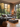 The image shows a modern living room with a large window showcasing autumn foliage. A gray sofa is adorned with various pillows and a throw blanket. The room also features a square gray coffee table with decorative items, wooden herringbone flooring, and a round woven basket. The scene creates a feeling of warmth and modern comfort.