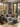 An inviting living room features a gray sofa with tan and brown pillows and a woven throw. A concrete coffee table with a cup and tray sits on a patterned rug. Large windows with black frames offer a view of autumn leaves outside, creating a warm and cozy atmosphere.