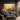 A stylish living room scene featuring a grey sofa with textured pillows and a patterned throw blanket, centered around a concrete coffee table with a wooden tray holding a French press and a mug. The focal point is a large black-framed window offering a view of vibrant autumn trees in full color. Natural light fills the room, highlighting the wooden floors and a large woven rug beneath the furniture.