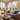 A bright and airy living room featuring a gray sofa with multiple pillows, a wooden coffee table with a cup of tea and a vase, and a wicker chair. Large windows fill the room with natural light, revealing an outdoor scene with trees displaying autumn colors. The overall mood is warm and inviting, suggesting a comfortable and stylish living space.