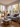 A bright and airy living room featuring a gray sofa with multiple pillows, a wooden coffee table with a cup of tea and a vase, and a wicker chair. Large windows fill the room with natural light, revealing an outdoor scene with trees displaying autumn colors. The overall mood is warm and inviting, suggesting a comfortable and stylish living space.
