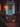 A warmly lit home entrance decorated for autumn and Halloween is shown. A blue door with a seasonal wreath is centered, flanked by a brick building with black shutters. The wooden steps are decorated with pumpkins, potted orange mums, and glowing lanterns containing candles, creating a cozy, inviting atmosphere.