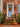 The image shows the front entrance of a brick home decorated for autumn. A weathered light blue door is centered in the frame, adorned with a colorful wreath made of fall leaves and berries. Several orange and white pumpkins of varying sizes, and pots of blooming mums line the brick steps, creating a festive and inviting scene.