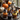 Several cupcakes with Halloween decorations are displayed on a silver tray. Each cupcake has a different color of frosting and is adorned with a small black witch hat. Pumpkins and a lit candle lantern are visible in the blurred background near a window, creating a warm autumn scene.