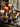 Several cupcakes with Halloween decorations are displayed on a silver tray. Each cupcake has a different color of frosting and is adorned with a small black witch hat. Pumpkins and a lit candle lantern are visible in the blurred background near a window, creating a warm autumn scene.