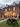 The exterior of a red brick house is decorated extensively for Halloween. A large black spider sits on the front lawn, with various skeletons, pumpkins, and other Halloween decorations arranged around the yard and on the front porch. The house's windows are illuminated from the inside, creating a warm and inviting, yet slightly spooky, ambiance.