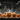 A chalkboard drawing depicts a Halloween night scene. At the bottom are various sized orange pumpkins with carved smiling faces. Above them are sketchy white drawings of houses and buildings forming a village, with lit up yellow windows. In the night sky, a crescent moon and a witch on a broomstick are sketched above the town.