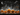 A chalkboard drawing depicts a Halloween night scene. At the bottom are various sized orange pumpkins with carved smiling faces. Above them are sketchy white drawings of houses and buildings forming a village, with lit up yellow windows. In the night sky, a crescent moon and a witch on a broomstick are sketched above the town.