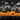 A whimsical illustration of a Halloween night scene. A row of smiling orange jack-o'-lanterns sits at the forefront. Behind the pumpkins are silhouetted spooky houses with glowing yellow windows, set against a dark night sky with white clouds, crescent moons, and twinkling stars. The overall effect is charming and spooky.