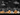 A chalkboard-style illustration depicts a Halloween scene. Several smiling jack-o'-lanterns of varying sizes are lined up in the foreground, with some pumpkins drawn in orange and others in white. Behind them is a row of small houses with simple outlines, set against a black night sky with a crescent moon and fluffy clouds.