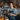 A young woman with freckles and blue eyes smiles directly at the viewer. She has her hair in a messy bun and is wearing a gray t-shirt and a denim apron with leather straps. She is holding a gray mug with latte art and is standing in a coffee shop with pumpkins on the counter in front of her and shelves filled with various items and dried flowers in the background.