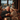 An elderly couple with gray hair embraces affectionately, both smiling broadly, as they sit by a large window. The man holds a warm mug, and the woman is wrapped in a cozy, patterned blanket, surrounded by soft candlelight and festive Christmas garland decorations. Outside the window, a blurred, snowy winter scene with distant lights is visible, emphasizing the warm and inviting indoor atmosphere.