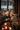 An elderly couple with gray hair embraces affectionately, both smiling broadly, as they sit by a large window. The man holds a warm mug, and the woman is wrapped in a cozy, patterned blanket, surrounded by soft candlelight and festive Christmas garland decorations. Outside the window, a blurred, snowy winter scene with distant lights is visible, emphasizing the warm and inviting indoor atmosphere.