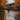 A couple walks away from the viewer down a residential street covered in fallen autumn leaves. Puddles of water reflect the gray sky. The trees lining the street are vibrant with orange and yellow leaves, framing the scene with seasonal color.