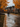 A couple walks away from the viewer down a residential street covered in fallen autumn leaves. Puddles of water reflect the gray sky. The trees lining the street are vibrant with orange and yellow leaves, framing the scene with seasonal color.