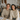 A smiling young man and woman in their 20s or early 30s sit side-by-side at a light wooden table, looking directly at the viewer. Both are wearing warm, textured knit sweaters in neutral shades, with the man in beige and the woman in cream. The background is a brightly lit, cozy room with a rustic aesthetic, featuring dried floral arrangements, string lights, and small autumnal decorations like pumpkins on the table alongside some papers.