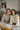 A smiling young man and woman in their 20s or early 30s sit side-by-side at a light wooden table, looking directly at the viewer. Both are wearing warm, textured knit sweaters in neutral shades, with the man in beige and the woman in cream. The background is a brightly lit, cozy room with a rustic aesthetic, featuring dried floral arrangements, string lights, and small autumnal decorations like pumpkins on the table alongside some papers.